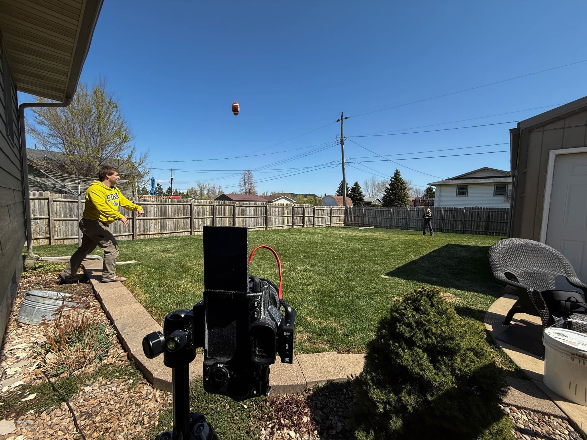 Real estate branding shoot in Whitewood, SD showing a photographer’s camera on a tripod capturing a man throwing a football across a backyard lawn beside a fenced house under clear skies.