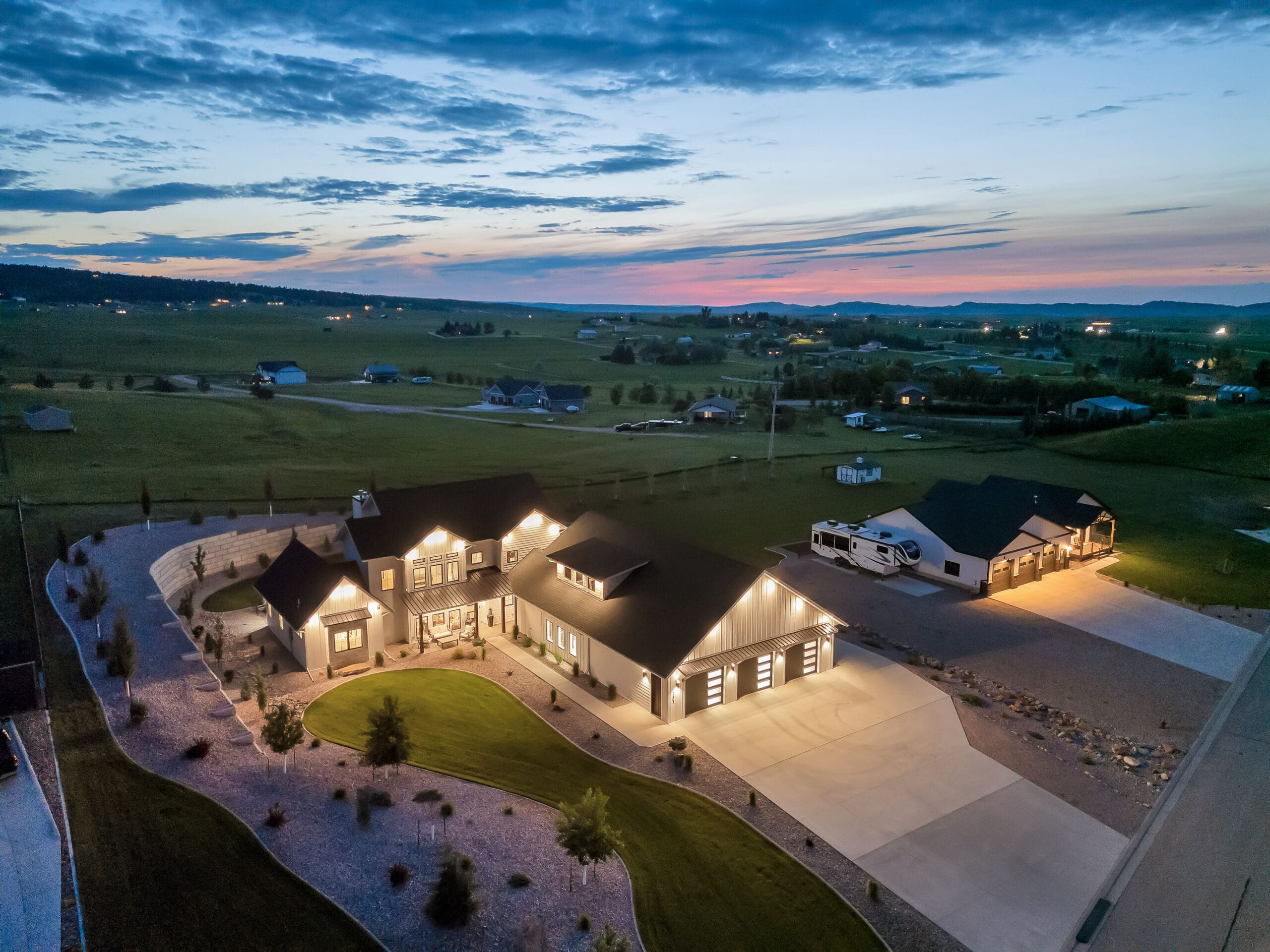 Drone shot of a luxury custom home and landscaped property at sunset in Spearfish, SD, captured by Odrix Media. The image showcases a well-lit house with a detached garage, RV parking, curved driveway, and scenic views of the surrounding rural neighborhood and hills.