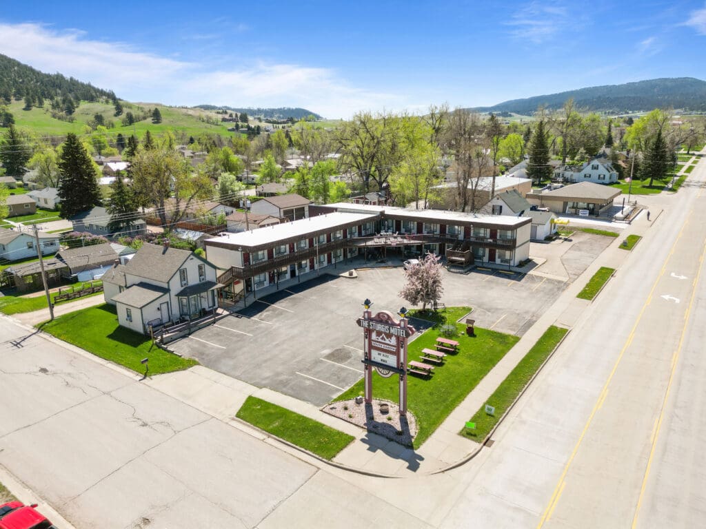 Aerial view of The Sturgis Motel captured by Odrix Media, showcasing the motel’s branding, U-shaped layout, parking area, and surrounding residential neighborhood in a small scenic town with green hills and trees in the background.