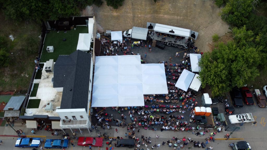 Drone aerial shot of a crowd gathered under white canopy tents in front of Mr. Wu’s building during the “Cool Deadwood Nights” concert event in Deadwood, South Dakota.