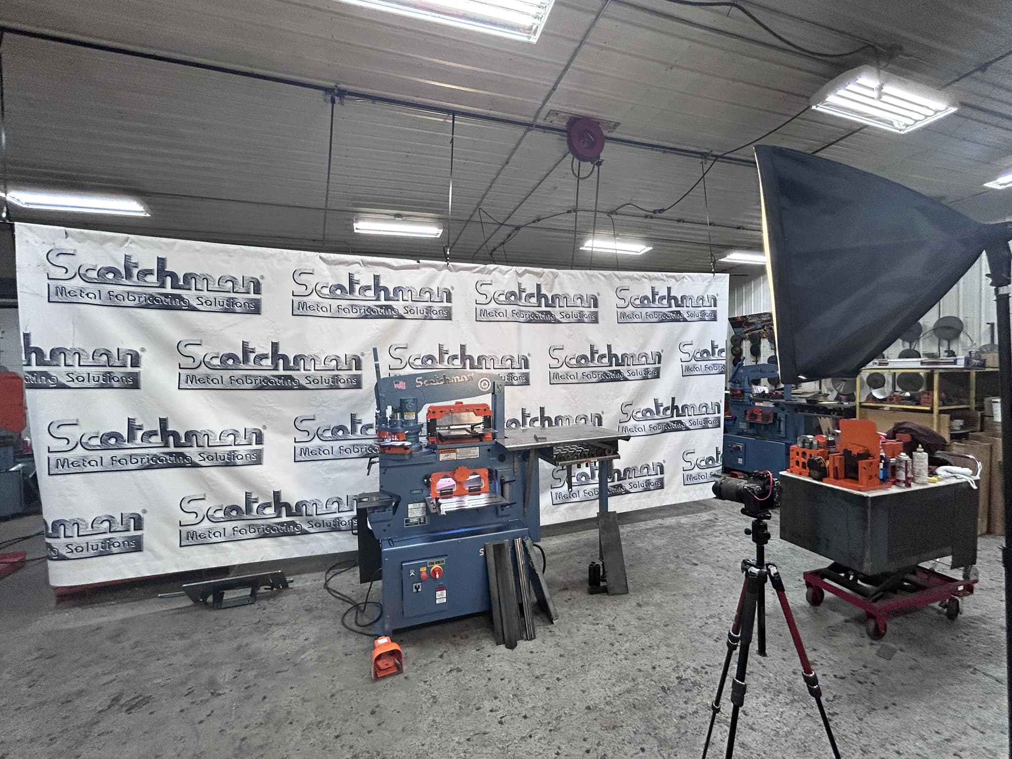 Workspace of a corporate branding shoot at Scotchman Industries in Philip, SD showing Scotchman metal-fabrication equipment set up against a branded backdrop, with a camera and softbox lighting positioned in front.