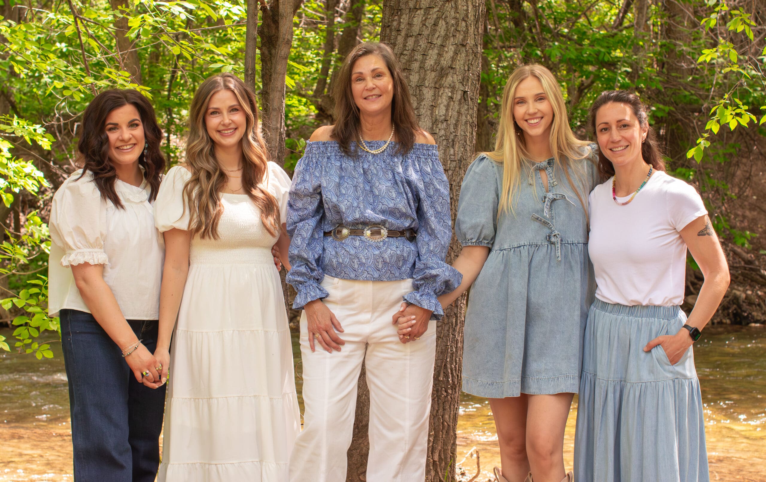 Family portrait session by Odrix Media and Genesis Ranch Photography featuring five women posing together outdoors along a wooded creek in the Black Hills of South Dakota.